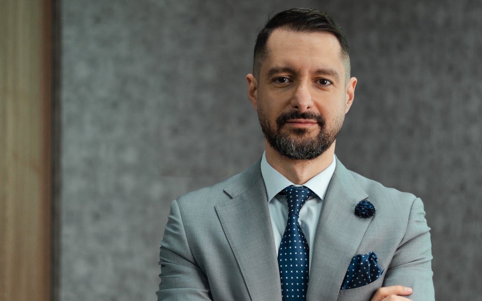 Portrait of Rafi Yachoua in a grey suit and navy polka-dot tie, arms crossed, against a concrete wall.